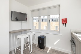 A room in Old Vicarage with a breakfast bar and stools. A television is mounted on the wall above the breakfast bar. Far wall has windows, a bin and a fire blanket secured to the wall.